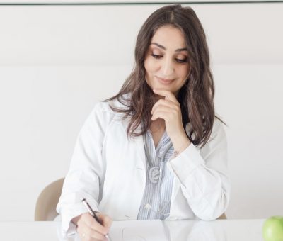 A female nutritionist in a lab coat writing notes with an apple and plant on the desk.